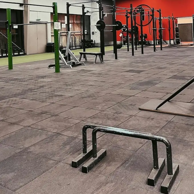 Espace de calisthénie en salle avec parallettes au premier plan, rigs et anneaux, sol en dalles caoutchouc, mur rouge.