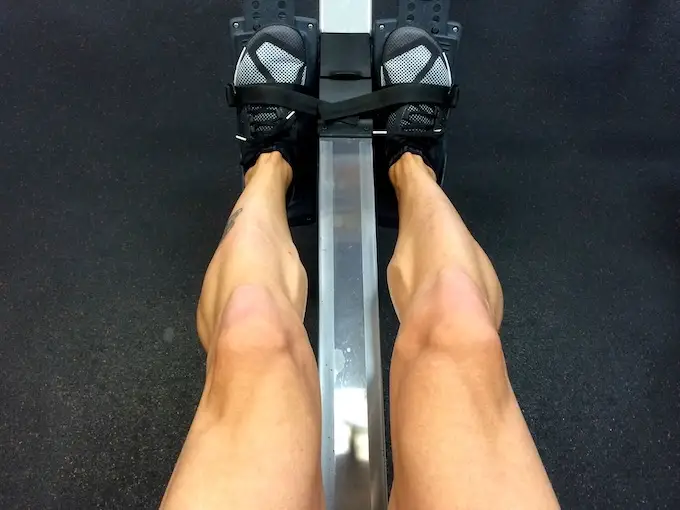 Vue du dessus des jambes sanglées sur les cale-pieds d’un rameur en salle de sport, chaussures noires, phase de poussée.