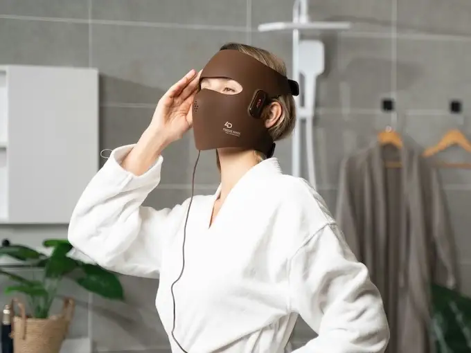 Femme en peignoir ajustant un masque LED facial marron dans une salle de bain moderne, soin beauté à domicile.