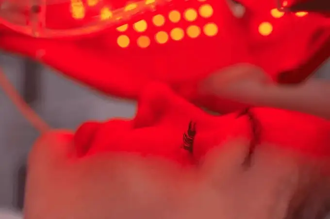 Visage en soin sous lumière rouge d’un masque LED, gros plan en institut, photobiomodulation esthétique, yeux fermés.