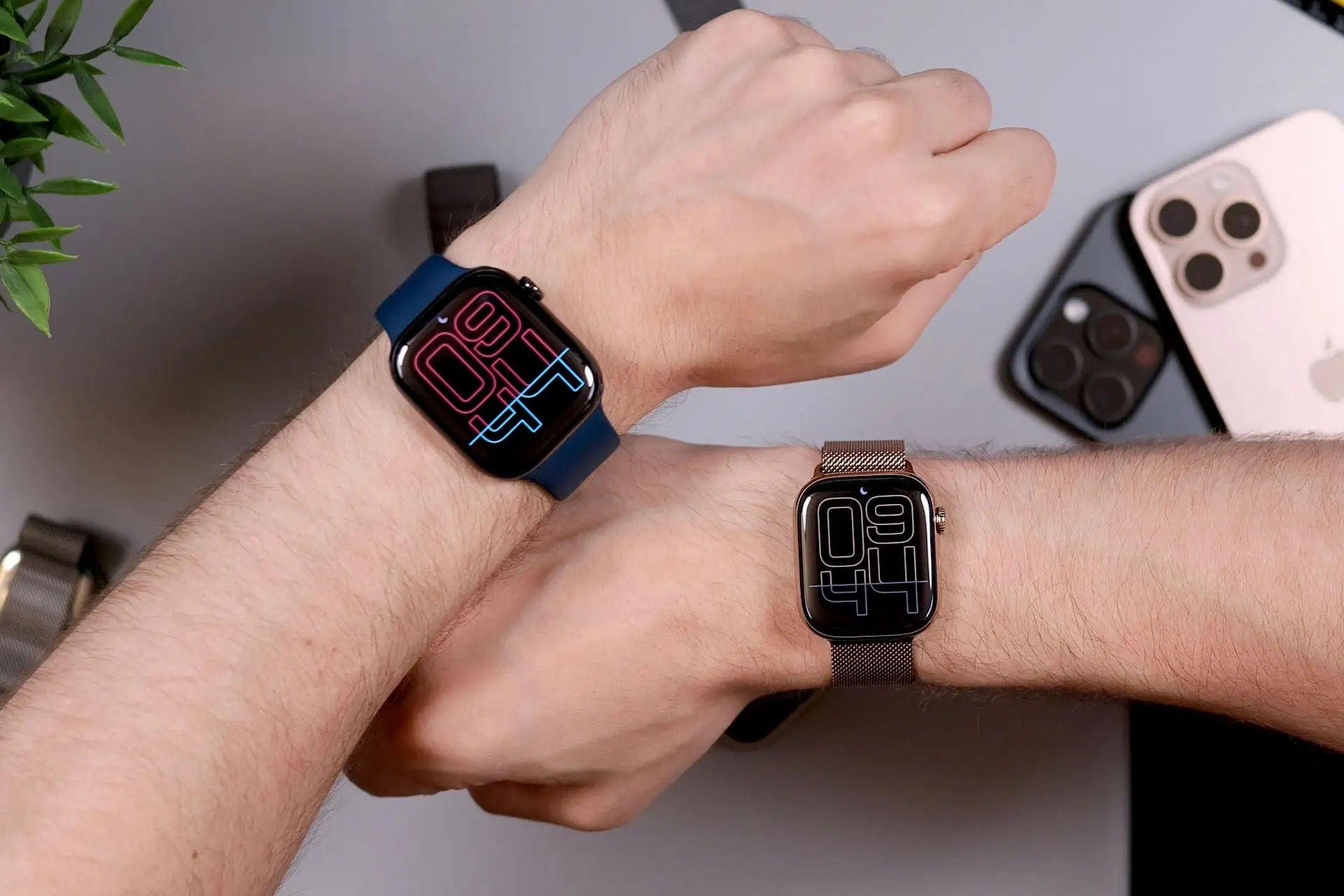 Deux montres connectées au poignet, cadran à grands chiffres néon, bracelets bleu et maille acier, smartphones en arrière-plan.