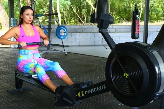 Sportive sur rameur Concept2 en salle, leggings multicolores, top rose, sac de frappe et disques en arrière-plan.